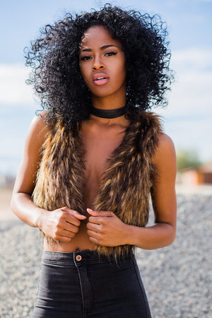 Attractive Black woman in a stylish fur vest poses outdoors against a blue sky. Her curly hair frames her face in this medium shot.
