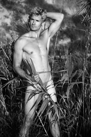 Black and white portrait of a shirtless adult man in a field, posing with tall grasses. He has fair hair, leans to the left, outdoors.