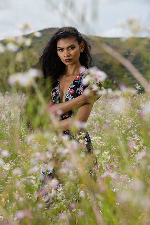 Woman in a floral dress stands in a field of white flowers with a mountain visible in the background, looking at the camera.