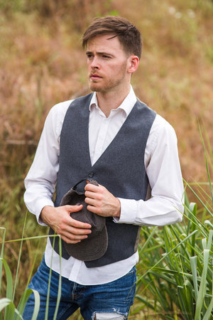 Portrait of a Caucasian male wearing a vest and jeans in an outdoor setting, holding a hat, with a field visible in the background.