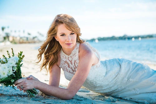 A woman in a white lace wedding dress is laying on the sand at the beach and holding a white floral bouquet, with ocean background.