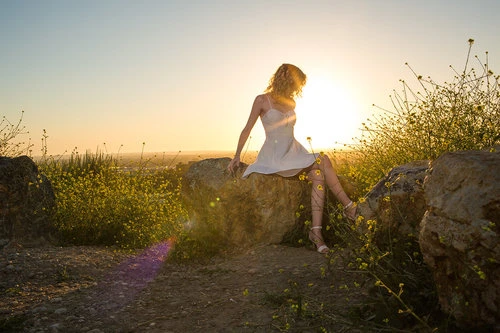 Woman in a white dress seated on large rocks surrounded by yellow wildflowers during a sunny sunset.