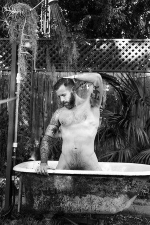 Black and white portrait of a bearded man in an outdoor bathtub under a shower, surrounded by lush foliage and a wooden fence.