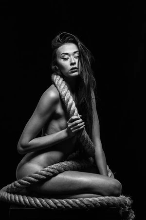 Black and white portrait of a woman seated with thick rope wrapped around her shoulders and body against a dark background.