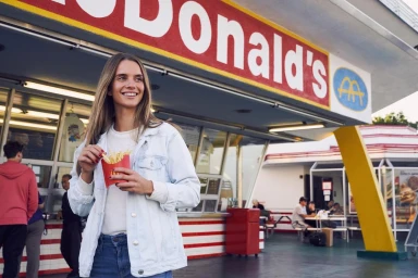 Smiling woman in light denim jacket eating french fries in front of a McDonald's restaurant exterior