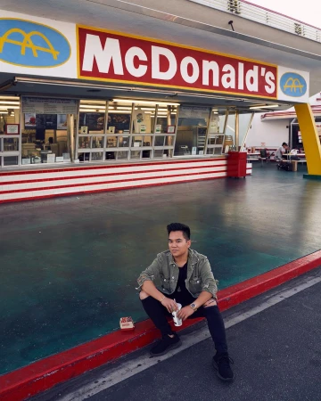 Man in distressed olive jacket and black ripped jeans sitting outside a McDonald's restaurant in Los Angeles by Alvin Nguyen.