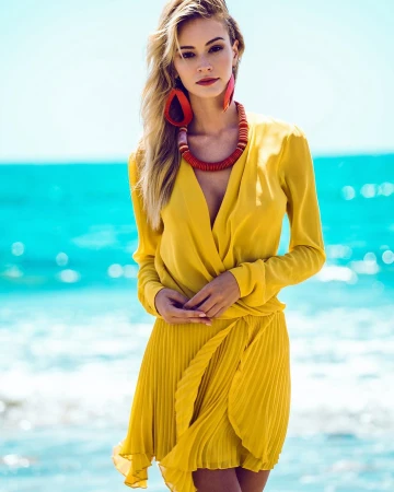 Model in a bright yellow dress with red drop earrings and a beaded necklace posing against an ocean background by Alvin Nguyen