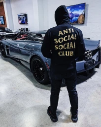 Man wearing a black hoodie with gold text standing next to a grey sports car in a showroom