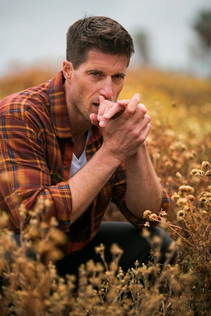Man crouching in a field of dry wildflowers wearing an orange plaid shirt with hands clasped near his face
