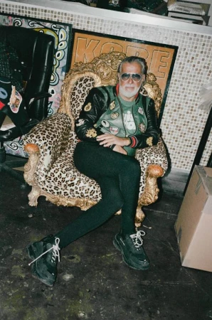 Elderly man with white beard and sunglasses seated in an ornate leopard print armchair wearing a patterned jacket and black trousers