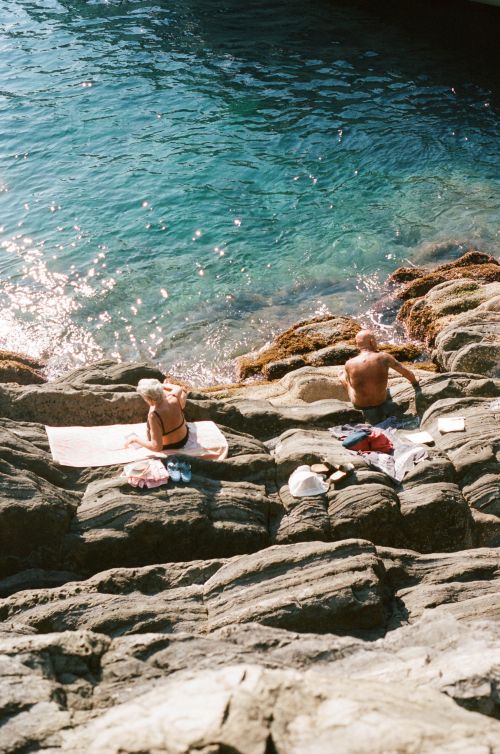 An elderly couple relaxing on a rocky beach by the turquoise sea; one sunbathing on a towel, the other sitting nearby with clothing items.