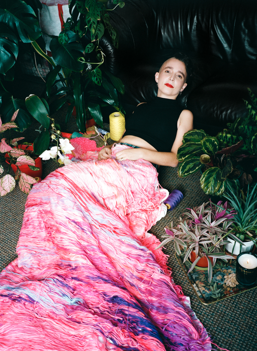 Adult woman lies on a rug surrounded by potted plants with a pink and purple fringed fabric draped over her in an indoor setting.