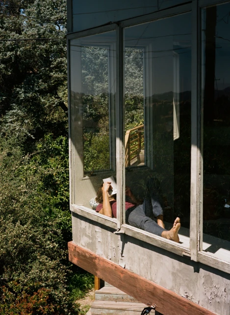 Adult relaxing indoors near a window with a book, enjoying natural light and a view of trees outside, creating a peaceful scene.