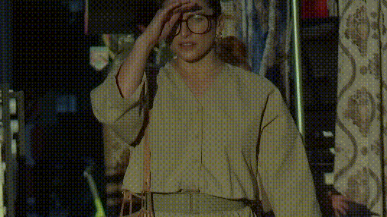 Woman in beige outfit with glasses adjusting her hair in a casual, sunlit indoor setting with patterned curtain in the background.