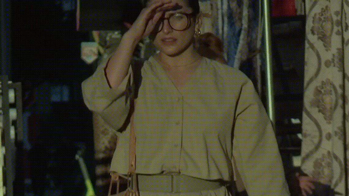 Woman in beige outfit with glasses adjusting her hair in a casual, sunlit indoor setting with patterned curtain in the background.