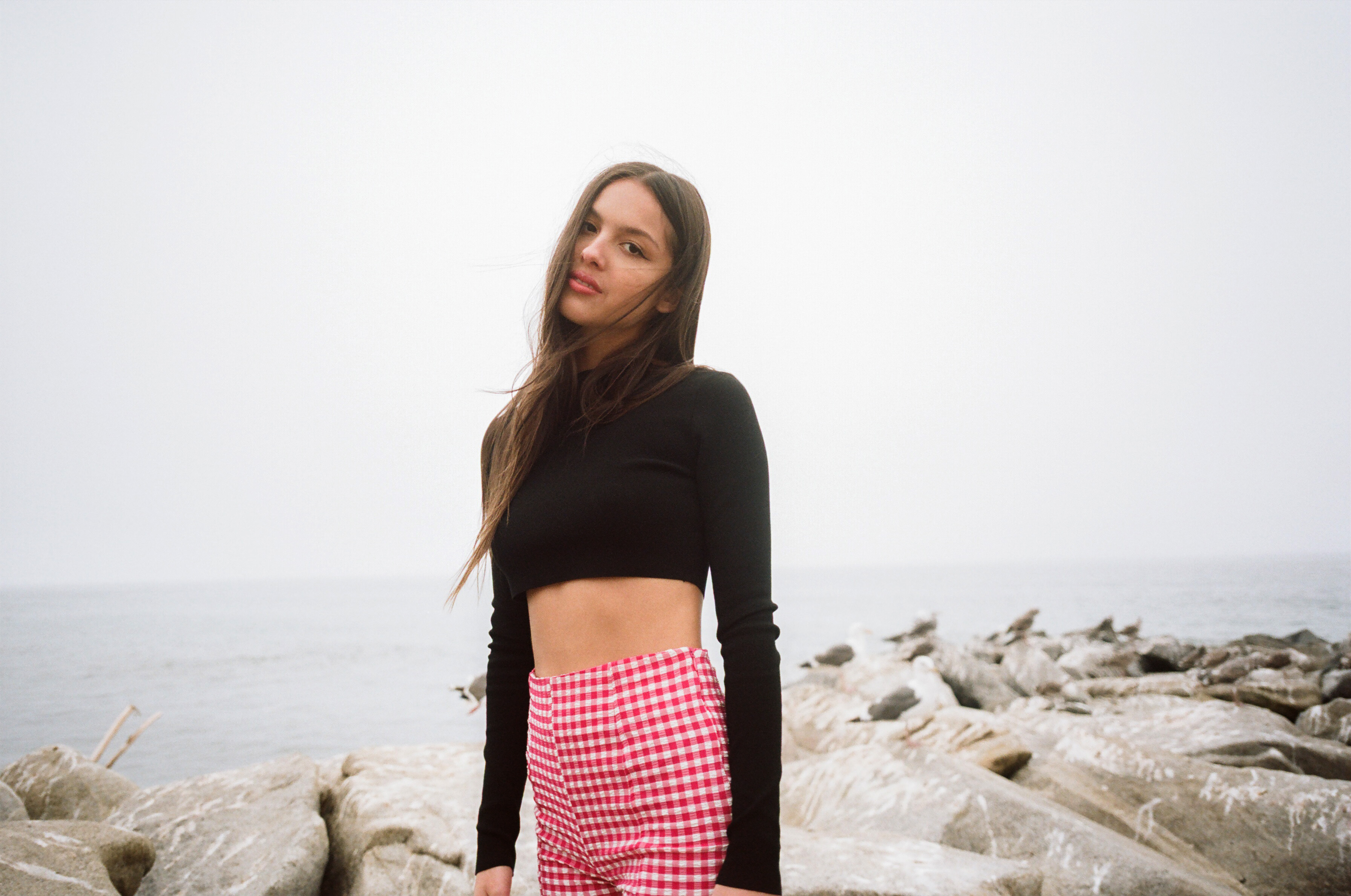 A young woman with long brown hair poses outdoors on a rocky shoreline wearing a black top and red gingham shorts against a misty ocean backdrop.