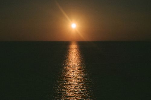 Golden hour at sea: A radiant sun shines over a dark, rippled ocean with a shimmering reflection trail, creating a serene seascape.