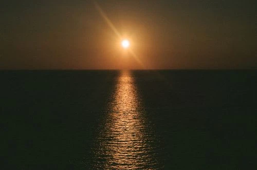Golden hour at sea: A radiant sun shines over a dark, rippled ocean with a shimmering reflection trail, creating a serene seascape.