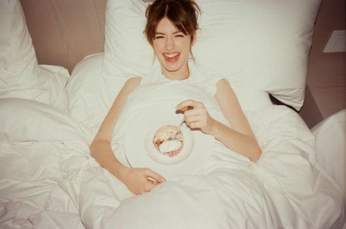 Young woman lies in bed eating dessert with a spoon, surrounded by white linens and pillows, showing a casual, relaxed lifestyle moment.