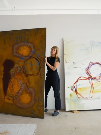 An artist stands in a bright studio next to their large, abstract paintings in brown and blue tones, creating a creative atmosphere.