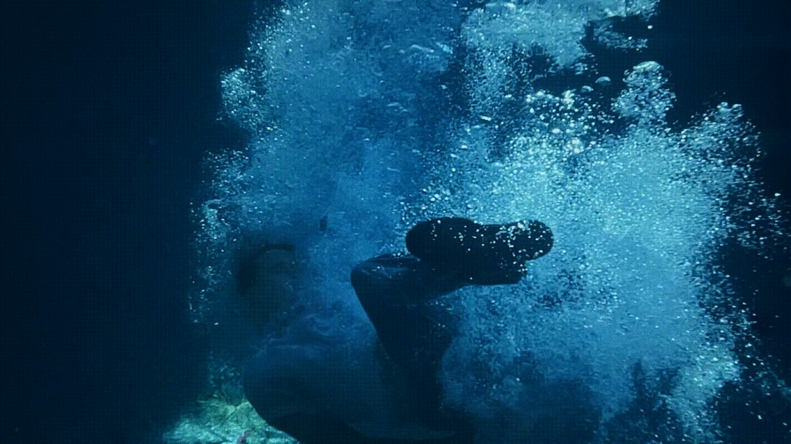 Person swimming underwater in blue ocean. Bubbles surround them. Underwater photography of person diving into water.