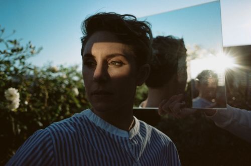Portrait of a woman in a blue and white striped shirt, holding a mirror reflecting a sunny outdoor scene with greenery.