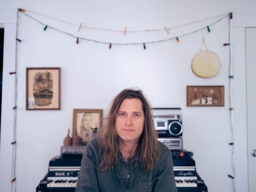 Portrait of an adult male musician with long hair sitting at an electric piano indoors in front of music equipment and string lights.
