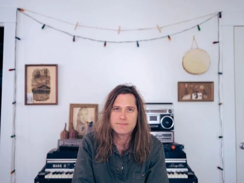 Portrait of an adult male musician with long hair sitting at an electric piano indoors in front of music equipment and string lights.
