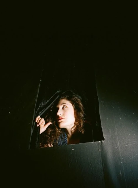 Woman with curly hair peers out from a dark enclosure, looking up and away with a neutral expression in a portrait shot.
