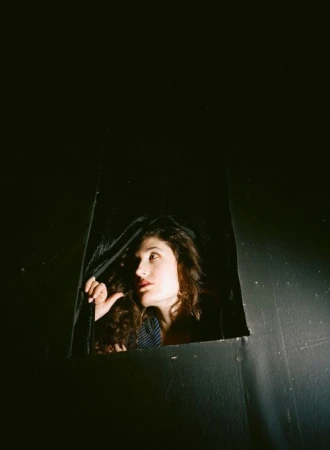 Woman with curly hair peers out from a dark enclosure, looking up and away with a neutral expression in a portrait shot.