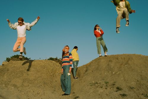 Group of four young adults in retro clothing posing on a hill against a blue sky, one jumping in the air.