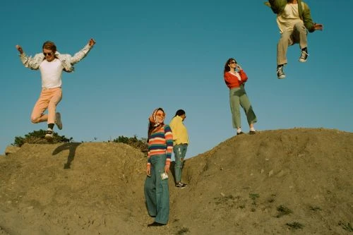 Group of four young adults in retro clothing posing on a hill against a blue sky, one jumping in the air.