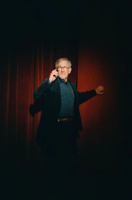 Adult male wearing glasses and a suit jacket gestures on a red stage in a dimly lit photo studio with a blurred background.