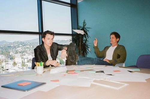 Two men in a modern office sitting at a table with paperwork, pens, laptop, and paper airplane, with a cityscape visible through large windows.