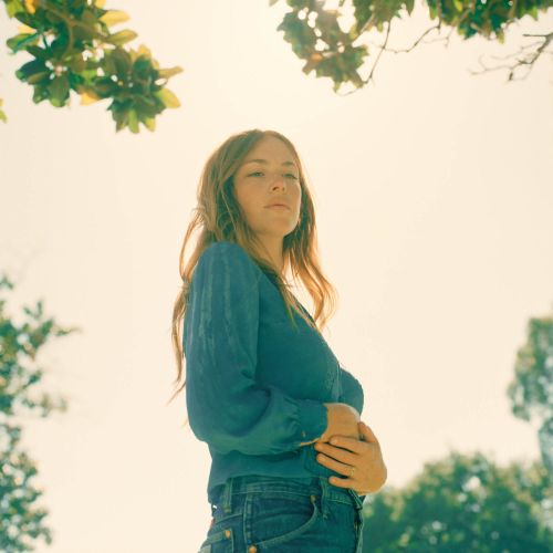 Portrait of a woman in jeans and a blue textured blouse, with sunlight and tree branches framing her against a bright sky.