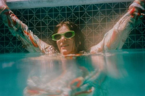 Woman submerged in a pool wearing sunglasses with hands raised, creating a cheerful summer vibe with turquoise water and geometric tile.