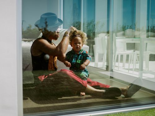 An adult wearing a blue hat styling a child's curly hair indoors near glass windows, captured by Chantal Anderson.