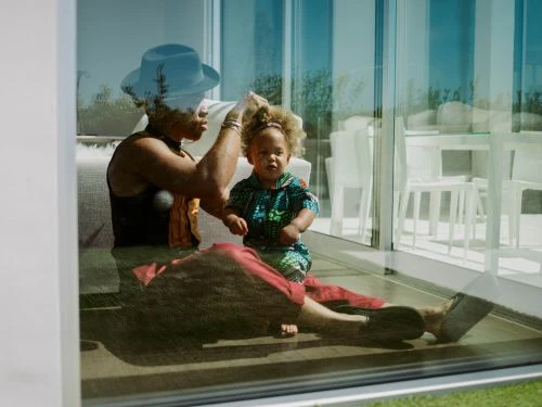 An adult wearing a blue hat styling a child's curly hair indoors near glass windows, captured by Chantal Anderson.
