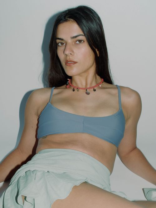 Young woman with dark hair and tanned skin wears a blue bra top and light-green skirt in a medium shot against a white background.