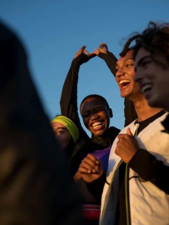 Group of young adults laughing outdoors against a blue sky, some wearing athletic apparel, showcasing a candid and joyful moment.