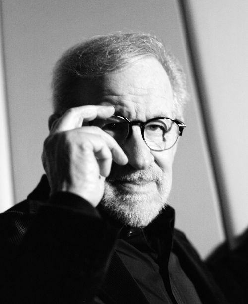Black and white portrait of an adult male with glasses, resting a finger on his forehead, in front of a neutral background.