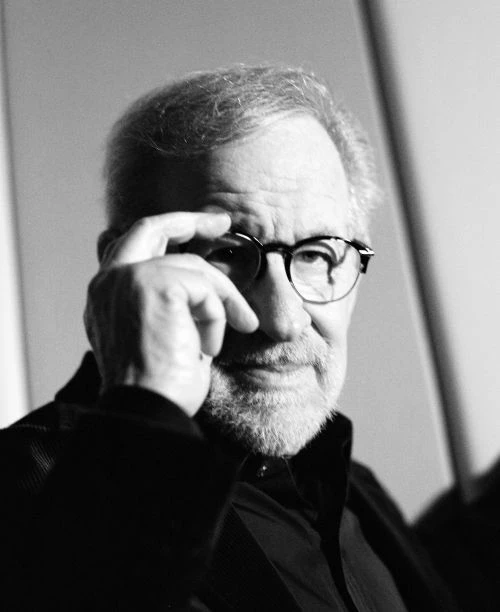 Black and white portrait of an adult male with glasses, resting a finger on his forehead, in front of a neutral background.