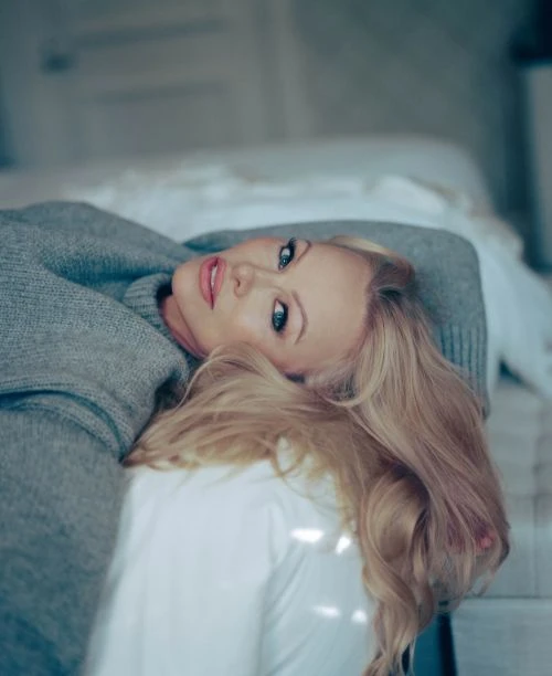 Blonde woman in a gray sweater lies on a white bed, looking up at the camera with a slight smile in an indoor setting.