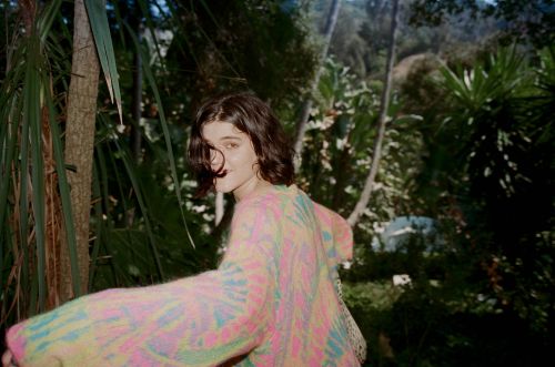 A young woman with short dark hair looks over her shoulder wearing a colorful tie-dye sweater in a lush tropical forest.