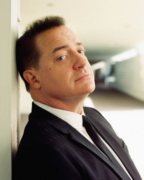 Portrait of Brendan Fraser in a black suit, posing against a white wall. The actor is seen looking at the camera with a slight smile.