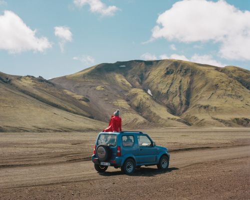Person in red jacket seated on roof of blue SUV parked on dirt track before large mountain landscape