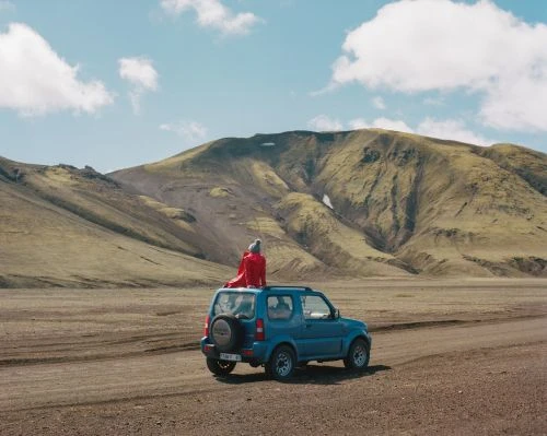 Person in red jacket seated on roof of blue SUV parked on dirt track before large mountain landscape