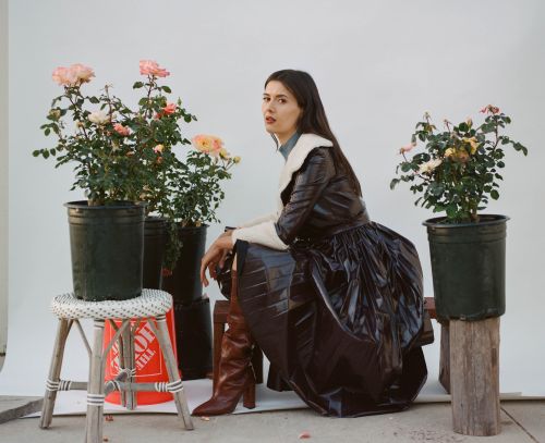 Portrait of a young woman in a dark, stylish coat sitting outside near rose bushes in pots against a white backdrop in soft light.