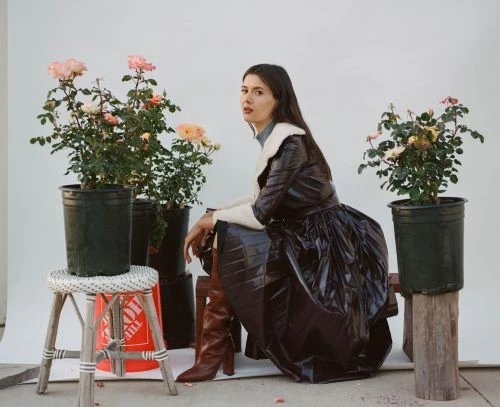 Portrait of a young woman in a dark, stylish coat sitting outside near rose bushes in pots against a white backdrop in soft light.