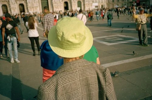 Crowd of people in a town square, featuring a person in the foreground wearing a bright green hat and plaid jacket.